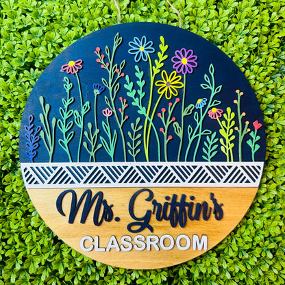 A round hanging classroom sign with a dark blue upper half featuring colorful layered flowers and green stems, a white geometric stripe, and a light wood lower half engraved with the words 'Ms. Griffin's CLASSROOM'.